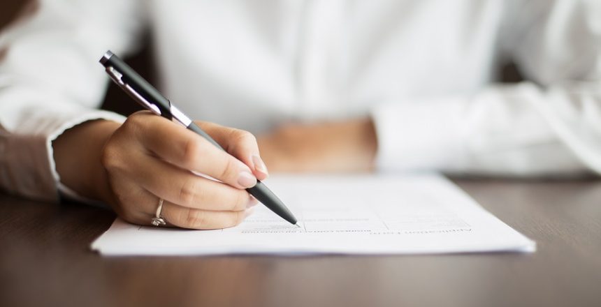 Hand of young businesswoman wearing ring and sitting at table writing on document with pen in office